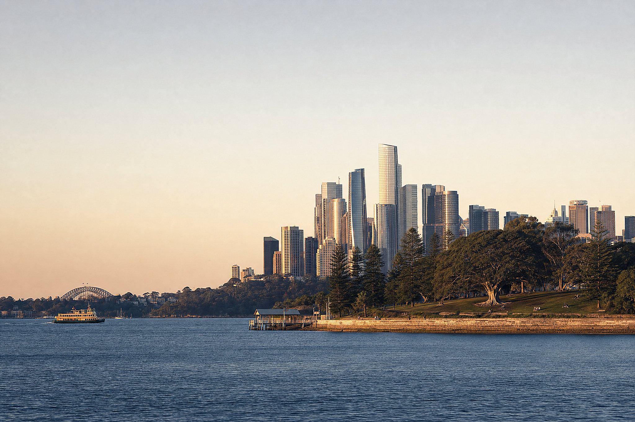 The Sydney skyline across the harbour at golden hour, the bridge and a ferry small on the water; at night, the towers lit